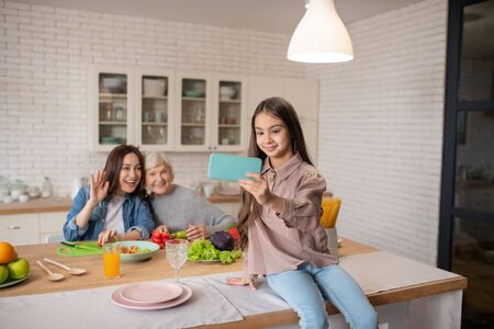 Good selfie. Joyful daughter sitting on a table in the kitchen taking a selfie with mom and...