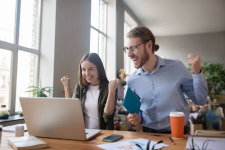 This is victory. Young enthusiastic girl and man in glasses and with a tablet in hand, standing in front of a laptop and looking at the screen, arms raised victoriously, smiling and happy.の写真素材