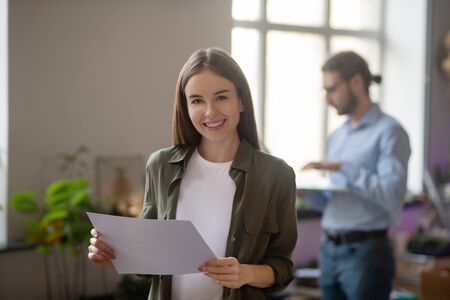 Favorite job. Smiling dark-haired long-haired girl in a good mood with a document in her hands, standing in the office and a man standing by the window.の写真素材