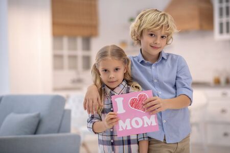 At home. Kids standing in the kitchen and holding sign, boy hugging girl, both smiling