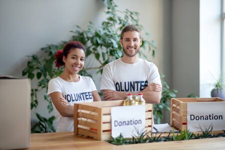 Work done by volunteers. Young man and girl in identical white T-shirts with the inscription volunteer standing in front of packed food boxes, arms folded across their chest smiling.の写真素材