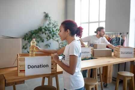 Caring for the needy. Young joyful girl in a white T-shirt putting a bottle of vegetable oil in a...
