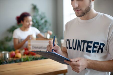 Summing up work. Young volunteer with beard with blue folder and pen in his hands, standing in room...
