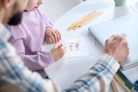 Drawing for dad. Little daughter in a pink sweater drawing a picture for dad with colored pencils,...