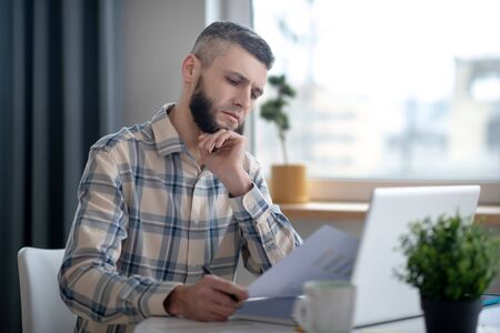 Work with charts. Young serious man sitting at a table in front of a laptop, holding a sheet of paper in his hand looking at him.の写真素材