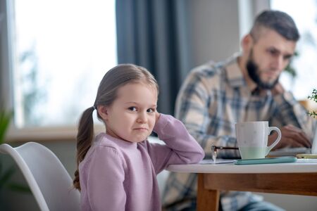 Sad daughter. Little cute girl in a pink blouse sitting at the table sadly smiling, dad nearby busy with work.の写真素材