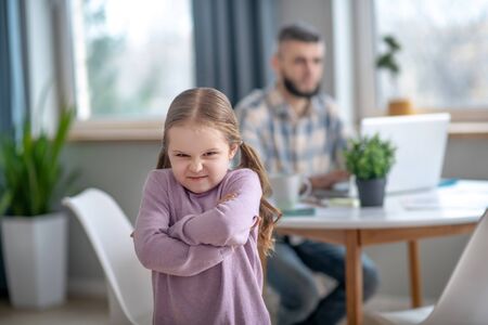 Resentment. Offended frowning little girl standing in the middle of the room with hands folded on her chest, dad busy sitting behind.の写真素材