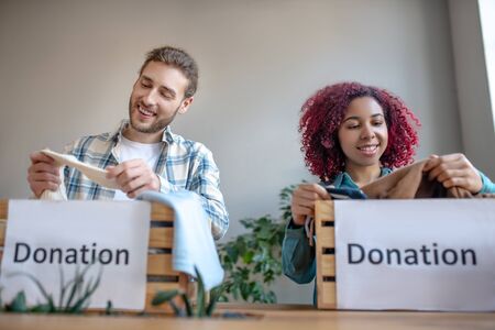 Charity sale. Smiling and happy young man and girl preparing clothes for a charity sale, pulling it out, examining and sorting.の写真素材