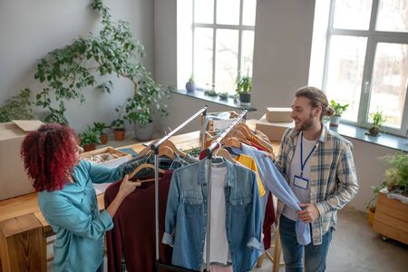 Hanging clothes. Young girl with curly hair and a man with a badge standing near a rack with clothes and sorting clothes on trempels for a charity sale, smiling cheerful.の写真素材