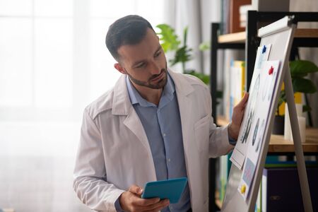 Needed drawings. Young bearded doctor in a white robe looking at the picture on a boardの写真素材