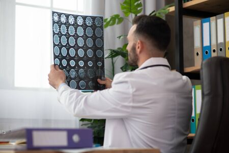 Neurologist at work. Young bearded doctor in a white robe working on the MRI results