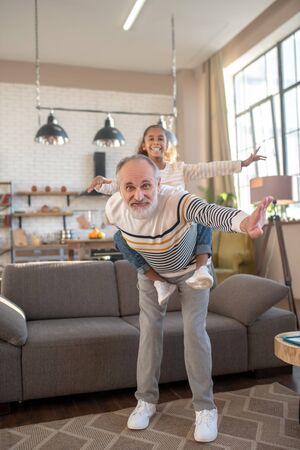 Weekend. Bearded grey-haired man holding his granddaughter on his backの写真素材