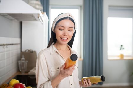 Healthy drink. Young asian woman standing in the kitchen with bottles of juice in her hands, looking at them smiling.の写真素材