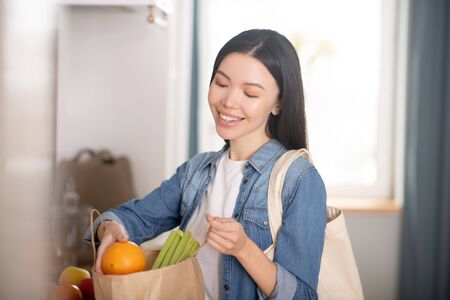Feeling good. Beautiful young woman with a paper bag of vegetables and fruits, pulling out an...