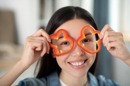 Life is beautiful. Dark-haired young girl holding slices of red pepper in her hands near her face...