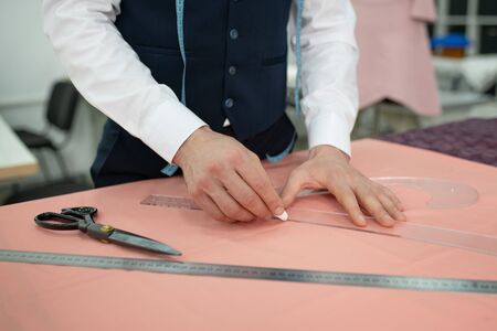 Fashion design studio. Hands of male tailor outlining pattern on pink cloth with chalk