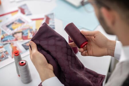 Fashion design studio. Male tailor holding dark scarlet cloth and spool, matching colors