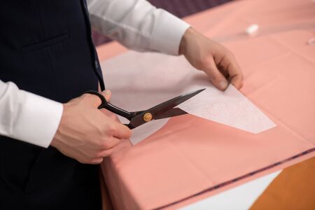 Fashion design studio. Hands of male tailor cutting out paper pattern over pink cloth