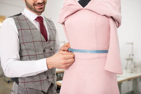 Fashion design studio. Hands of male tailor measuring waist of pink dress with blue measuring tape