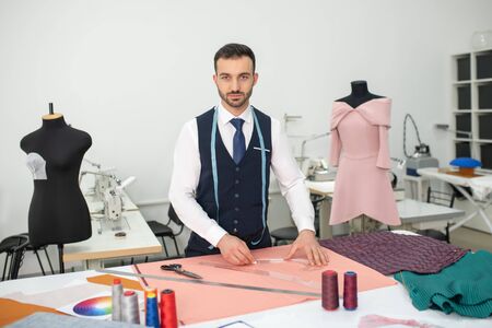 Fashion design studio. Male tailor standing and outlining pattern on pink cloth, looking into the...