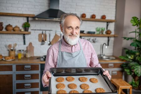 Cooking skills. A smiling pensioner wearing apron holding just baked cookiesの写真素材