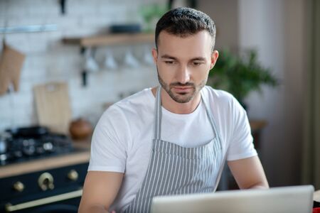 Search for a recipe. Successful dark-haired young man in a white T-shirt and striped apron looking...