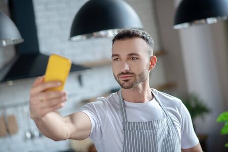 Selfies. Young man in a white T-shirt and apron, standing at home with a yellow smartphone in his hand, taking a selfie, serious and satisfied.の写真素材