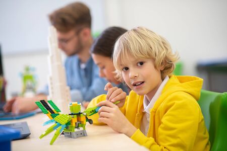 School classroom. Blond boy playing with buildable bird, dark-haired girl and male teacher sitting...