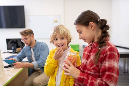 School classroom. Kids eating sweets and smiling, male teacher holding tablet and scrolling