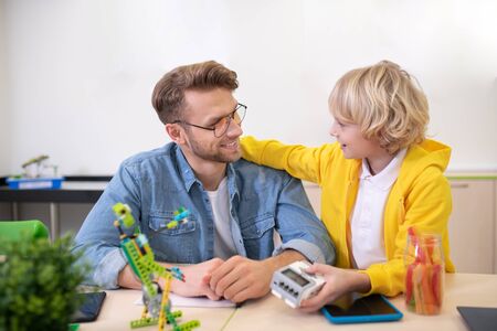 School classroom. Blond boy holding control unit, embracing male teacher, both smiling