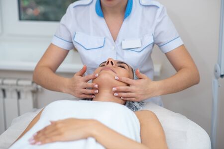 Doing face massage. Beauty doctor wearing uniform doing face massage for client lying and enjoyingの写真素材