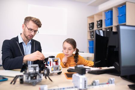 School classroom. Male teacher and dark-haired girl sitting at desk, he fixing buildable car, dark-haired girl touching her chin, feeling interested and involvedの写真素材