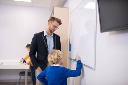 School classroom. Male teacher standing at whiteboard with hands in pockets, boy writing something,...