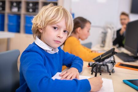 School classroom. Blond boy and dark-haired girl sitting at the desk, taking notes, male teacher sitting at the computer, looking at themの写真素材