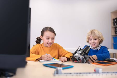 School classroom. Pupils sitting at the desk, taking notes, playing with buildable car, smiling, feeling excitedの写真素材
