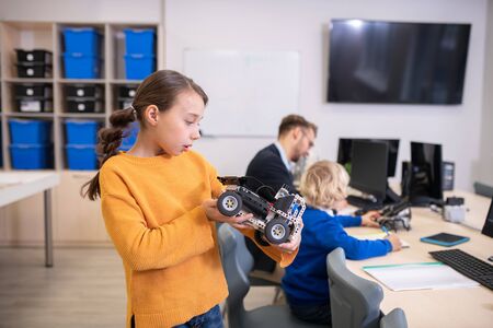 School classroom. Dark-haired girl holding buildable car, looking carefully at it, male teacher and...