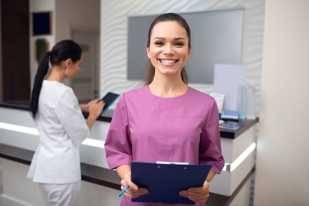 Smiling broadly. Beaming woman wearing uniform working in beauty clinic smiling broadlyの写真素材