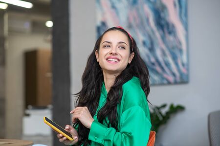 Communication. Woman with long dark thick hair looking joyfully to the side, holding a smartphone in...