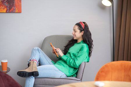 Online. One woman looking at a tablet while sitting in a soft chair, in a good cheerful mood.