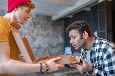 Smelling good. Dark-haired man in a checkered shirt smelling pizza from a boxの写真素材