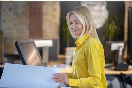 At workshop. Blonde woman standing at printing machine, holding paper, smiling