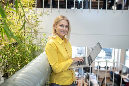 At work. Blonde woman standing on stair landing, holding laptop, typing, smiling
