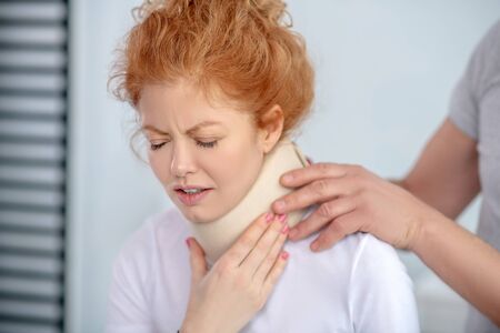 At rehabilitation center. Male physiotherapist hands examining neck fixator, female patient frowning from painの写真素材
