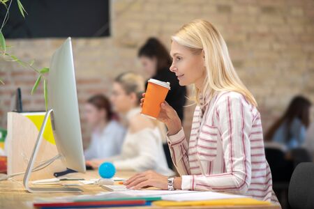 Modern office. Blonde woman working at computer, drinking coffee