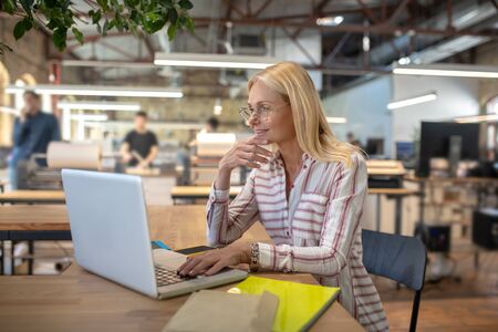 At work. Blonde woman sitting at laptop, checking something, holding her chinの写真素材