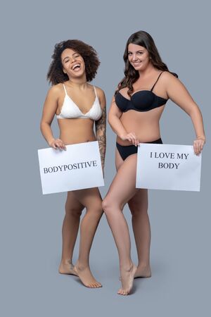 Health, beauty, body. Beautiful young women in white and black lingerie standing near barefoot, foot on toe, holding down posters in their hands, rejoicing laughing.の写真素材