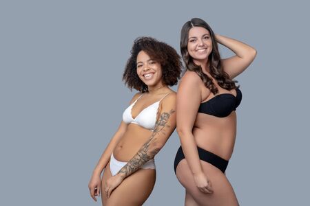 Not ashamed, body. Woman with curly hair and jewelry on the neck in white lingerie, her long-haired friend with curvaceous in black, standing with their backs to each other, rejoicing.の写真素材