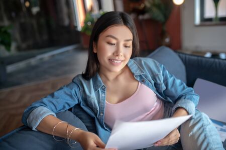At home. Young woman sitting on sofa, holding papers, reading, smilingの写真素材