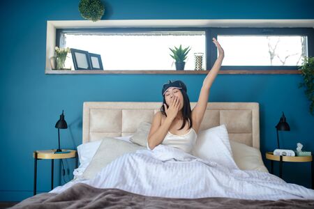 At home. Young woman in sleeping eye mask yawning and stretching in bedの写真素材