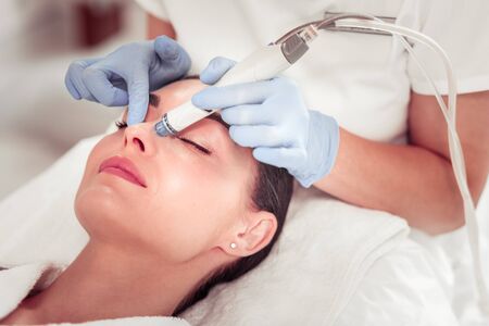 Using modern equipment. Cosmetologist wearing gloves using modern equipment for face cleansing